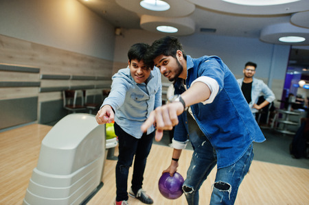 South asian mans in jeans shirt standing at bowling alley with ball on hands and aiming. Guy is...