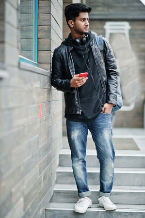 Stylish and casual asian man in black leather jacket, headphones with red mobile phone at hands posed on street.の写真素材