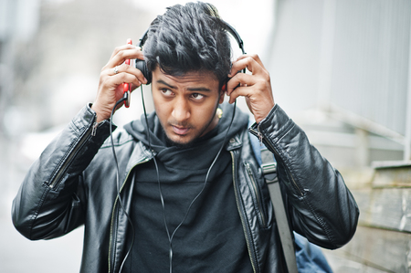 Stylish and casual asian man in black leather jacket, headphones with red mobile phone at hands posed on street and listening music.の写真素材