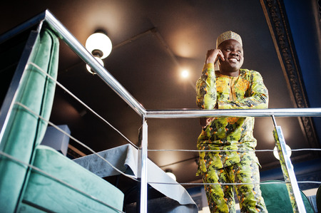 Friendly afro man in traditional yellow clothes with cap at restaurant near the handrail speaking at...