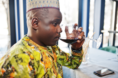 Friendly afro man in traditional yellow clothes and cap drinking red wine at restaurant.の写真素材