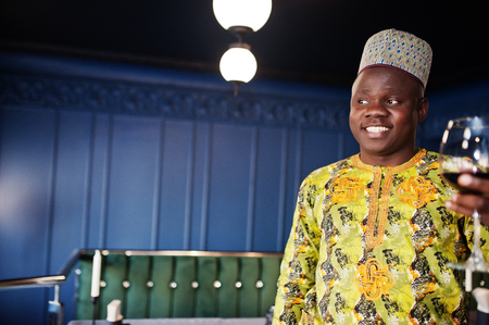 Friendly afro man in traditional yellow clothes and cap with red wine at glass in restaurant.の写真素材