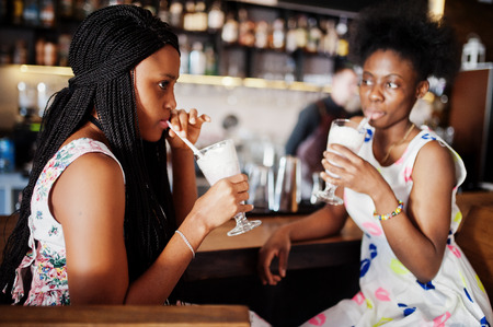 Two black african girlfriends at summer dresses drinking milkshake cocktails in bar.の写真素材
