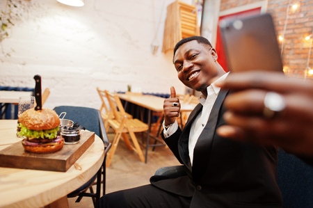 Respectable young african american man in black suit sitting in restaurant and making selfie against tasty double burger and show thumb up.の写真素材