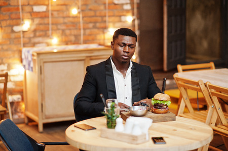 Respectable young african american man in black suit sitting in restaurant with tasty double burger and soda drink.の写真素材