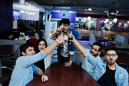 Group of stylish asian friends wear on jeans sitting at table and cheering with soda bottles in...