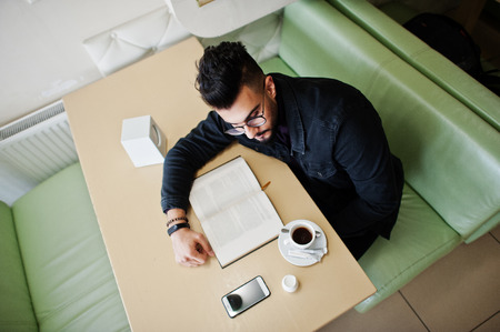 Arab man wear on black jeans jacket and eyeglasses sitting in cafe, read book and drink coffee.の写真素材