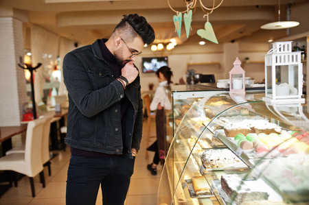 Arab man wear on black jeans jacket and eyeglasses in cafe choose cakes.