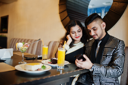 Lovely indian couple in love, wear at saree and elegant suit, sitting on restaurant and looking at phone together. On table dessert cakes, ice cream and juice.の写真素材