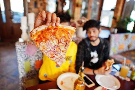 Asian friends guys eating pizza during party at pizzeria. Happy indian people having fun together, eating italian food and sitting on couch.の写真素材