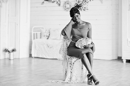 Fashion african american model in red beauty dress, sexy woman posing evening gown sitting at chair in white vintage room.の写真素材