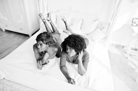 Two fashion african american models in red beauty dress and golden high heels, sexy womans posing evening gown lie on the bed.の写真素材