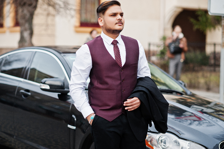 Stylish indian businessman in formal wear vest suit standing against black business car on street of city.の写真素材