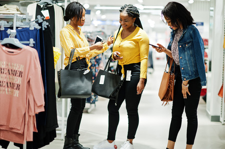 Three african woman at clothes store with new handbags. Shopping day. They look at mobile phones.の写真素材