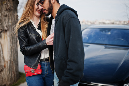 Cool multiracial couple hugs together against black elegant sport car.