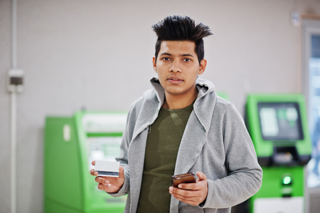 Young stylish asian man with mobile phone and credit card in hands against row of green ATM.の写真素材