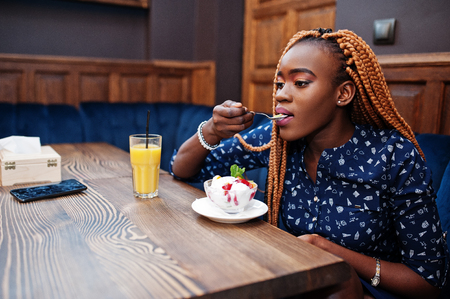 Portrait of beautiful young african business woman with dreadlocks, wear on blue blouse and skirt, sitting in cafe with ice cream and pineapple juice.の写真素材