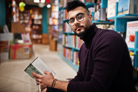Tall smart arab student man, wear on violet turtleneck and eyeglasses, at library sitting against books shelves.の写真素材