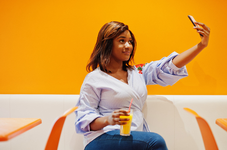 African woman sitting on cafe against orange wall with pineapple juice in hands and making selfie.の写真素材