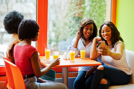 Four young african girls with juices sitting in bright colored fast food restaurant and making selfie by phone.の写真素材