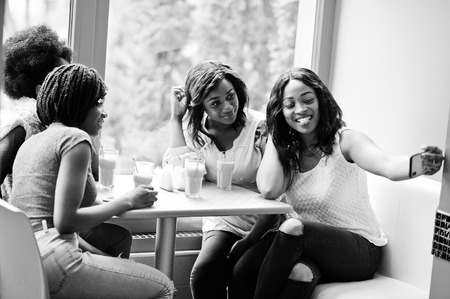 Four young african girls with juices sitting in bright colored fast food restaurant and making selfie by phone.の写真素材