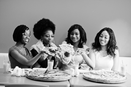 Four young african girls in bright colored restaurant holding pizza slices in hands.の写真素材