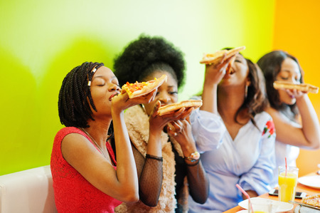 Four young african girls in bright colored restaurant eating pizza slices in hands.の写真素材