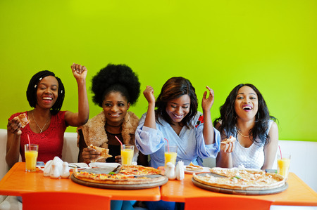 Four young african girls in bright colored restaurant eating pizza and having fun together.の写真素材