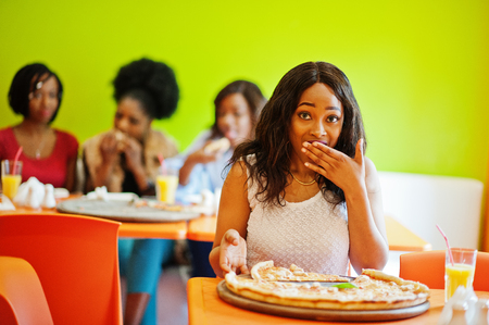 African woman with pizza sitting at restaurant against dark skinned girls. Surprised face.の写真素材