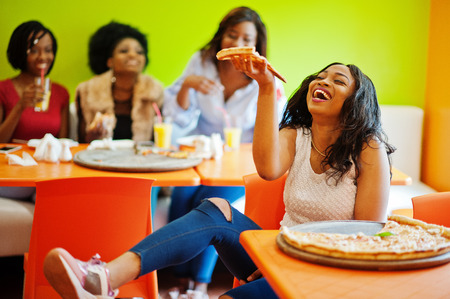 African woman with pizza sitting at restaurant against dark skinned girls.の写真素材