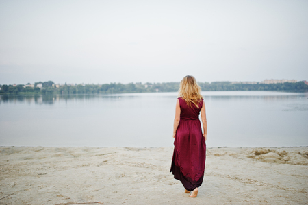 Back of blonde sensual barefoot woman in red marsala dress posing against lake.の写真素材