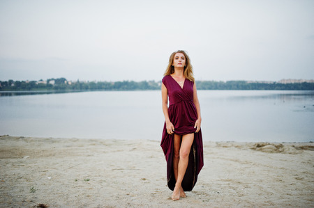 Blonde sensual barefoot woman in red marsala dress posing against lake on sand.の写真素材