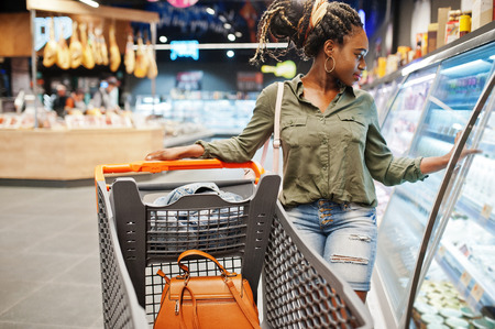 African woman with shopping cart choose yogurt bottle from fridge at supermarket.の写真素材