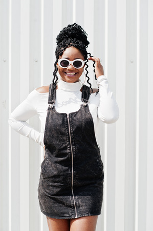 African woman in denim overalls skirt, white sunglasses posed against white steel wall.の写真素材