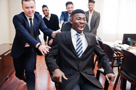 Six multiracial business mans standing at office and roll man on chair. Diverse group of male employees in formal wear having fun.の写真素材