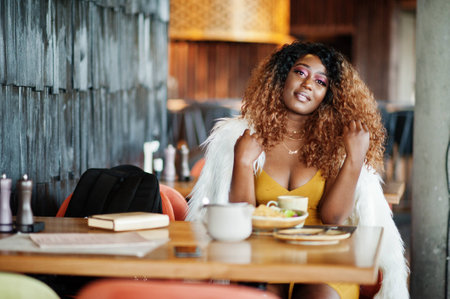 Glamour african american woman in yellow dress sitting at the table with dishes on restaurant.の写真素材