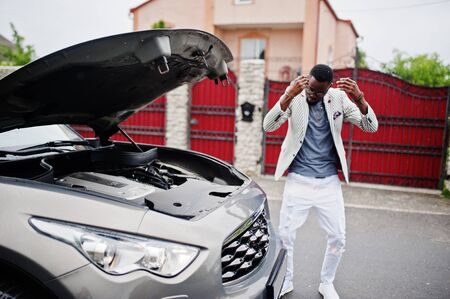 Stylish and rich african american man stand in front of a broken suv car needs assistance looking under opened hood.の写真素材