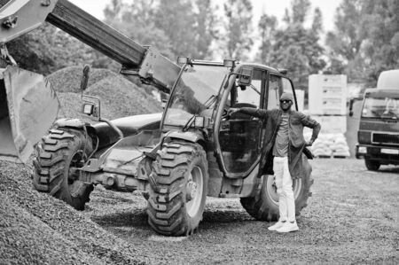 Stylish african american man in hat and sunglasses posed outdoor in rain against tractor with a bucket.の写真素材