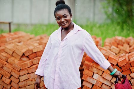 African woman in pink large shirt posed against bricks.