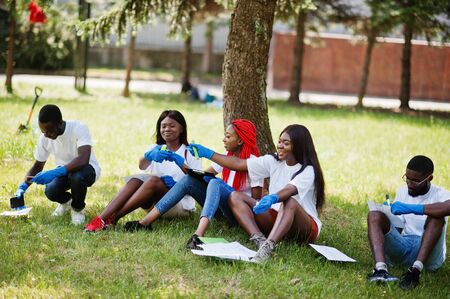 Group of happy african volunteers sitting under tree at park and write something on clipboards. Africa volunteering, charity, people and ecology concept.の写真素材