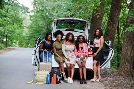 Group of five happy african american traveler girls sitting in car open trunk.の写真素材