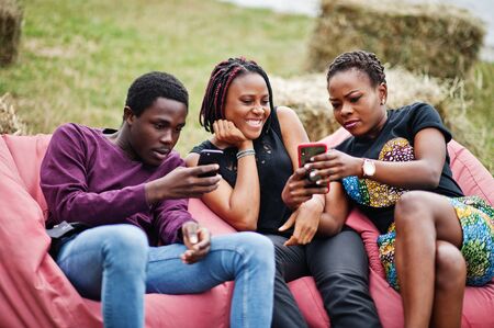 Three african american friends chill, sitting on poufs and using their phones outdoor.の写真素材