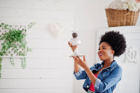 Attractive african american woman with afro hair wear on skirt and jeans jacket, posed at white room. Fashionable black model hold old gasoline lamp.の写真素材