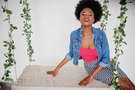 Attractive african american woman with afro hair wear on skirt and jeans jacket, posed at white room...