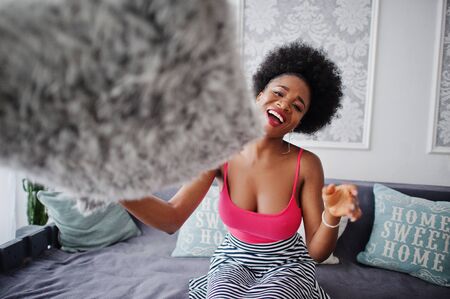 Attractive african american woman with afro hair wear on skirt pink top, posed at white room. Fashionable sexy black model play with pillows.の写真素材
