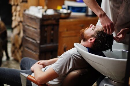 Young bearded man washing head by hairdresser while sitting in chair at barbershop. Barber soul.の写真素材