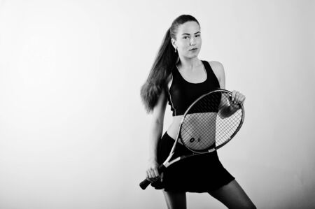 Black and white portrait of beautiful young woman player in sports clothes holding tennis racket while standing against whiteの写真素材