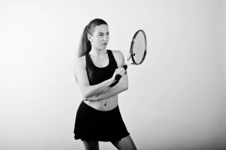 Black and white portrait of beautiful young woman player in sports clothes holding tennis racket while standing against whiteの写真素材