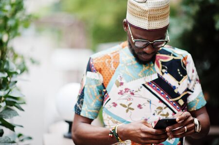 Handsome afro american man wearing traditional clothes, cap and eyeglasses in modern city looking at his cell phone.の写真素材