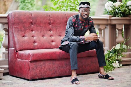 Handsome afro american man wearing traditional clothes, cap and eyeglasses in modern city sitting at couch.の写真素材